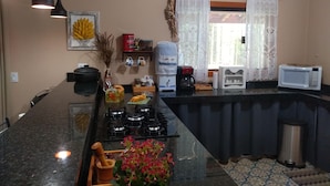 A kitchen with a stove, microwave, and coffee maker. There are hanging lights, a window with curtains, and a vase with flowers on the counter. - Sopro da Serra (Gonçalves)