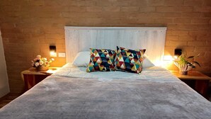 A bed with a grey blanket and colorful geometric pillows, a wooden headboard, and bedside tables with lamps and small plants. - Sopro da Serra (Gonçalves)