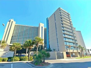 Een modern hotel met meerdere verdiepingen, een plat dak en een parkeerplaats met palmbomen.