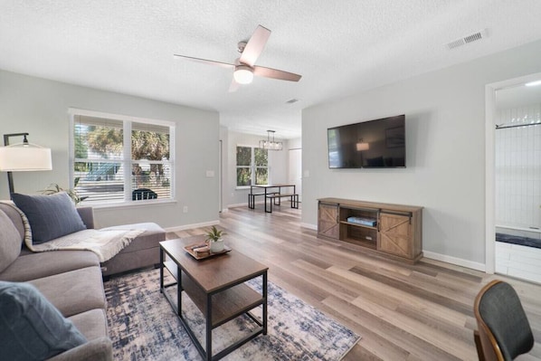 A spacious living room with a sofa, coffee table, and fireplace. There is a ceiling fan, a TV mounted on the wall, and large windows offering a view of the outdoors. - NEW Modern Bungalow | Close to Downtown! | Fenced Yard | Pet Friendly! (St. Augustine)