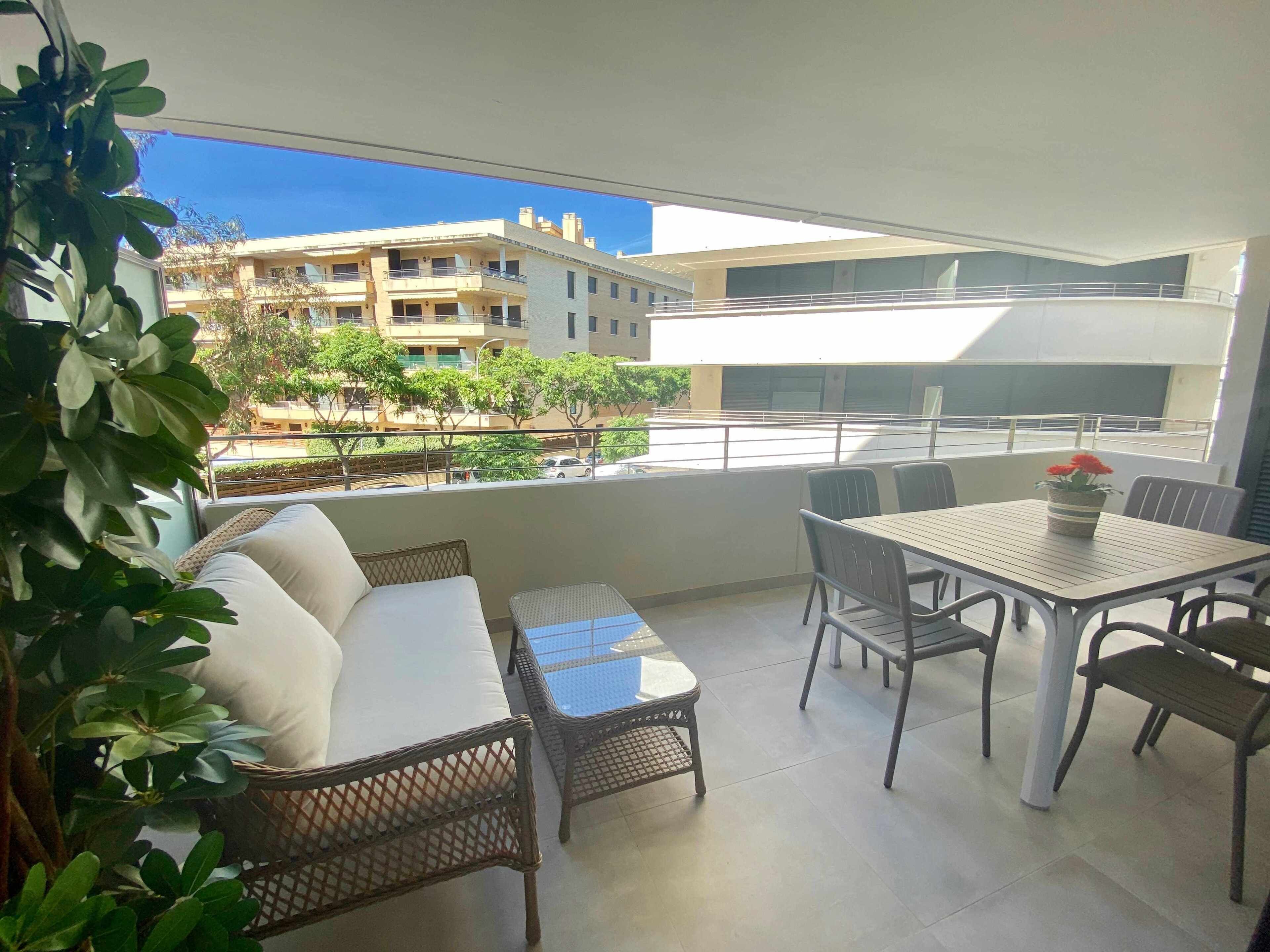 A balcony with a sofa, a small table, and chairs. There is a view of buildings and trees in the background.