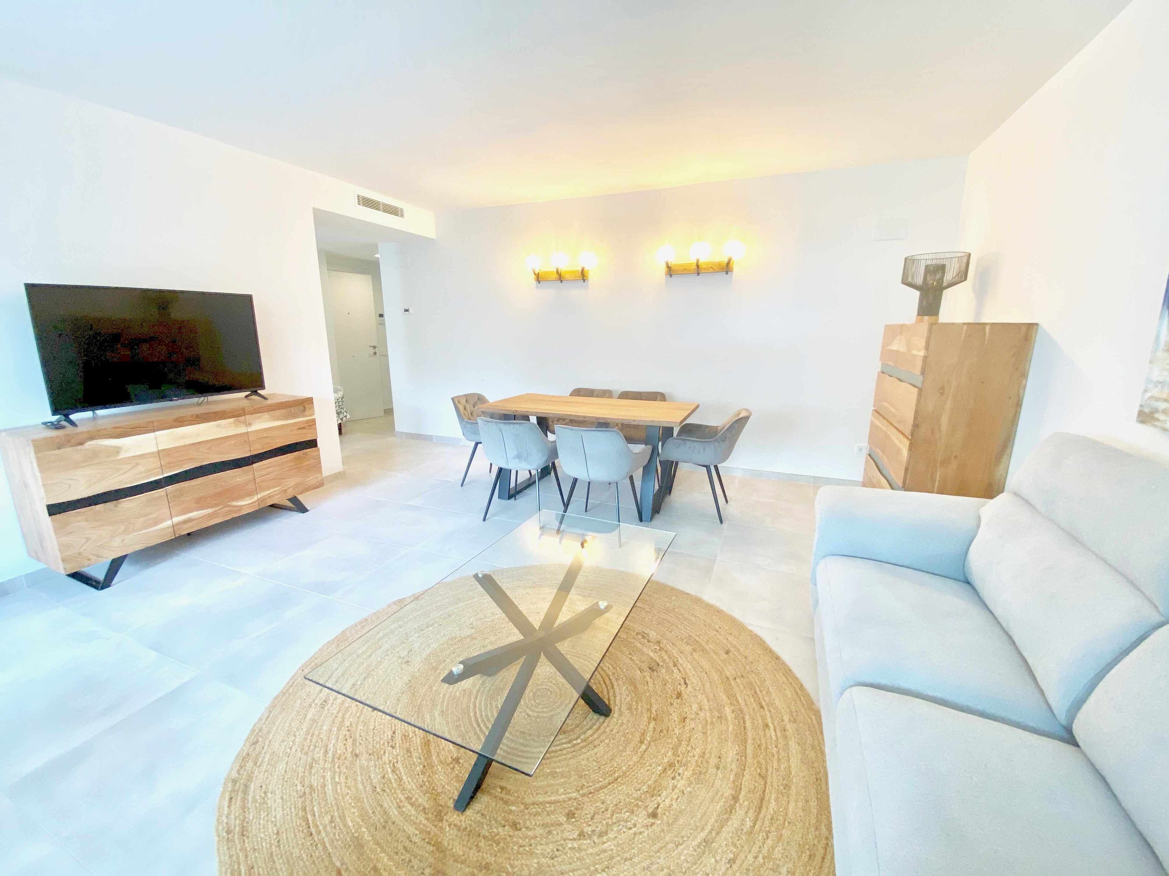 Una sala de estar moderna con televisor, mueble bajo de madera, mesa de comedor con sillas y un sofá de color claro.