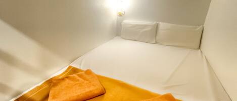 A small, white, single-bed room with a bright overhead light, white walls, and a yellow blanket on the bed.