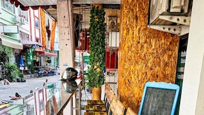 Filea di sedie e tavoli di legno in una zona esterna con pavimento piastrellato, una pianta in vaso e una lavagna.