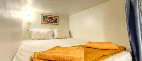 Un lit simple avec du linge de lit blanc et deux serviettes orange pliées, un tableau coloré accroché au mur et un luminaire au-dessus du lit.