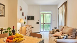 A living room with a sofa, a dining table with fruit, a TV, and a door leading outside.