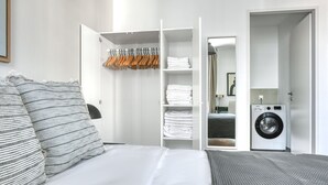 A hotel room with a bed, a wardrobe with hangers, a folded towel shelf, a washing machine, and a door leading to another room. - Sunny Leopoldstadt 1BR w/ Gym & Sauna, near U2, by Blueground (Vienna)