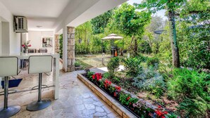 A covered outdoor area with a stone floor, two white bar stools, a small garden with red flowers, and a gazebo.