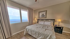 Ein Schlafzimmer mit einem großen Bett, zwei Nachttischlampen, einem Nachttisch, einem gerahmten Bild an der Wand und Meerblick durch das Fenster.