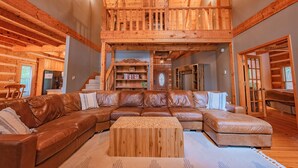 A spacious living room with a large brown leather sectional sofa, a wooden coffee table, and a wooden staircase leading to an upper level. - Burnt Mountain Waterfront Log Cabin, with 6person Spa  (Big Canoe)