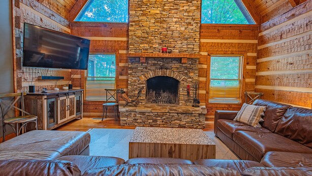 A log cabin interior with a stone fireplace, leather sofas, a flat-screen TV, and wooden walls. - Burnt Mountain Waterfront Log Cabin, with 6person Spa  (Big Canoe)