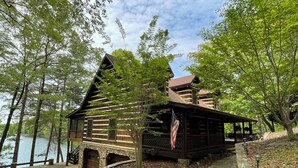 A log cabin with a stone staircase leading up to a wooden deck, surrounded by trees and a body of water. - Burnt Mountain Waterfront Log Cabin, with 6person Spa  (Big Canoe)