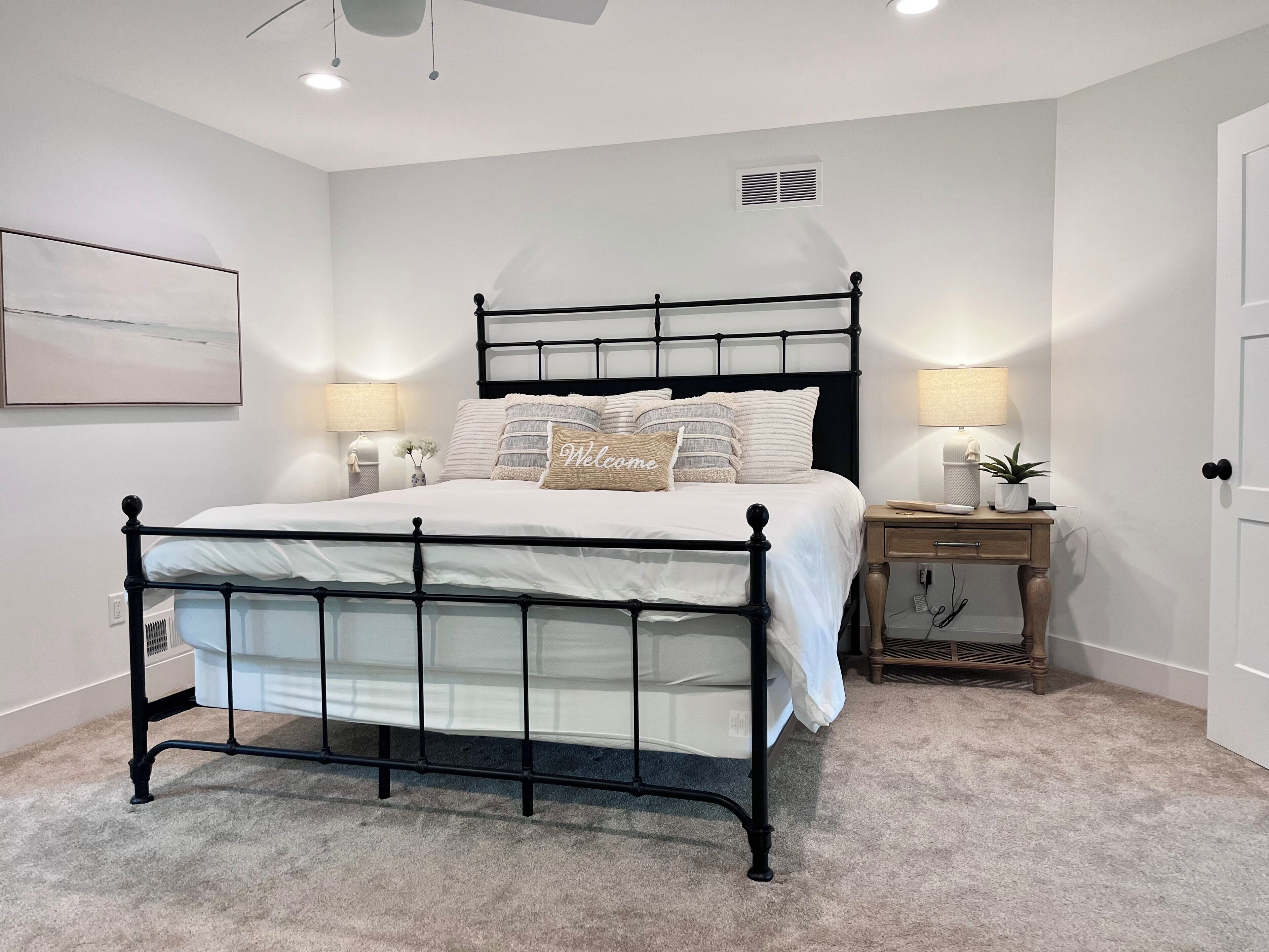 Une chambre à coucher avec un lit en métal noir, du linge de lit blanc, deux tables de chevet avec des lampes, une petite plante en pot et une bouche d’aération fixée au mur.