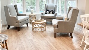 A spacious living room with large windows, a rattan coffee table, and a grey armchair with a pillow that says "OJUMP O LAKE".