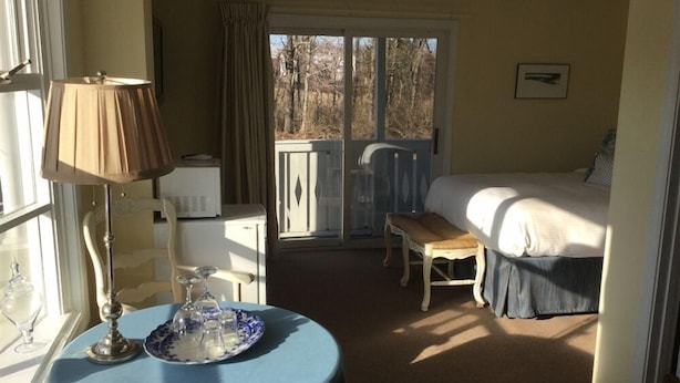 A hotel room with a bed, a table with a lamp, a chair, a balcony with a view of trees, and a small table with a plate and glasses. - The Beach Rose Inn (Wells)