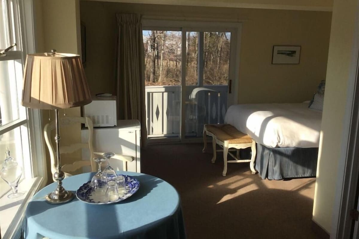 A hotel room with a bed, a table with a lamp, a chair, and a view through a sliding door to the outside.