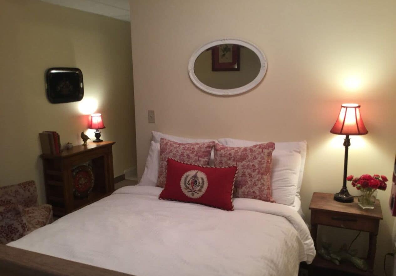 A neatly made bed with a red pillow, two bedside lamps, a small table with a vase of flowers, and a mirror on the wall.