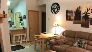 Un salon avec un canapé marron, une table basse en bois, une table à manger avec des chaises, un ventilateur de plafond et des œuvres d’art murales.