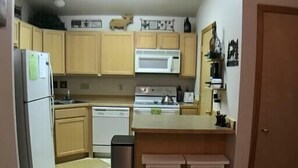 Une cuisine compacte avec des meubles en bois, un réfrigérateur blanc, un four à micro-ondes, une cuisinière et un lave-vaisselle. Il y a deux tabourets au comptoir et une poubelle.