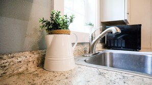 Une cuisine dotée d’un plan de travail en granit, d’un pichet blanc orné d’une plante, d’un évier en acier inoxydable et d’un micro-ondes noir.
