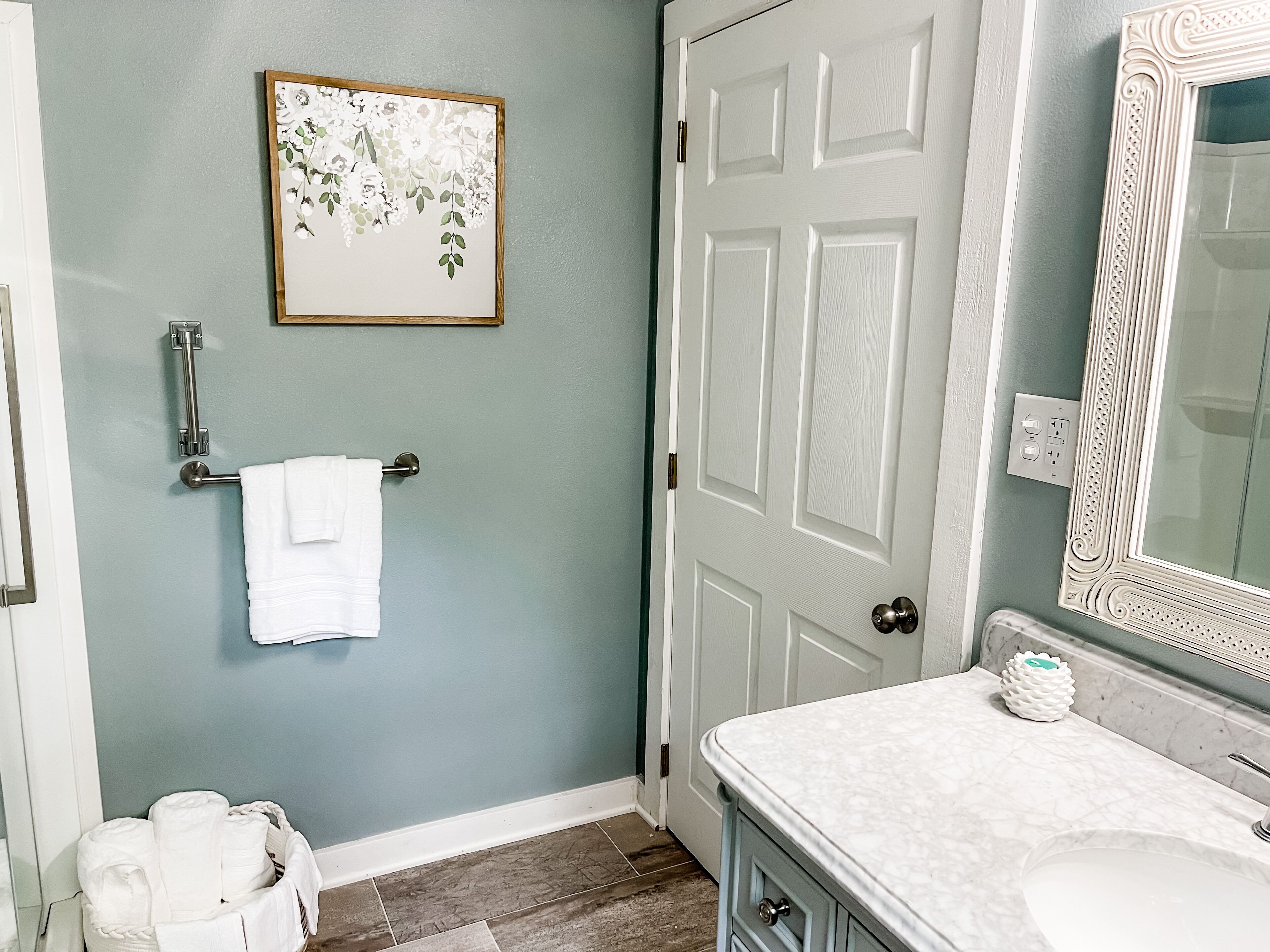 A bathroom with a white door, a framed floral artwork on the wall, a towel rack, a white bathrobe, a white sink, and a marble countertop.