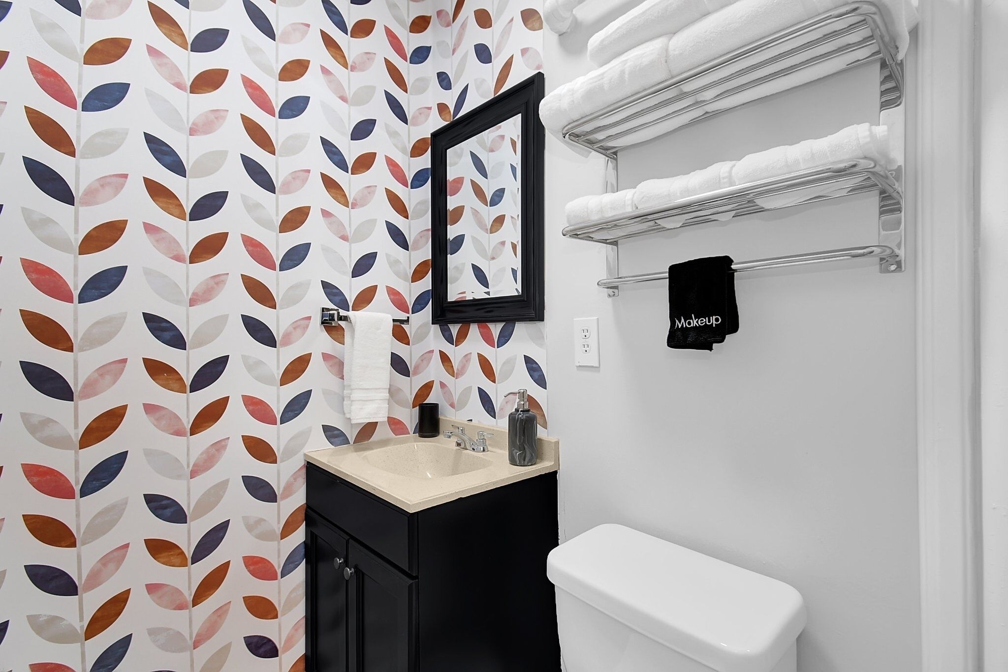 Une salle de bain avec des toilettes blanches, un lavabo avec un plan de toilette beige et un miroir au-dessus. On y trouve un porte-serviettes avec des serviettes blanches, ainsi qu’une serviette noire qui y est accrochée. Le mur est recouvert d’un papier peint à motifs de feuilles.