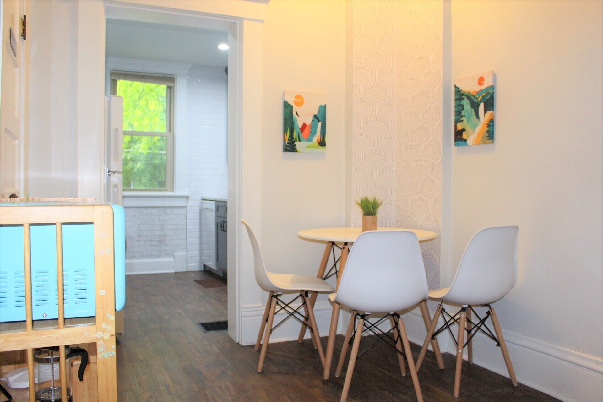 A modern kitchen with a dining area, featuring a wooden cabinet, a small table with chairs, and two wall-mounted artworks.