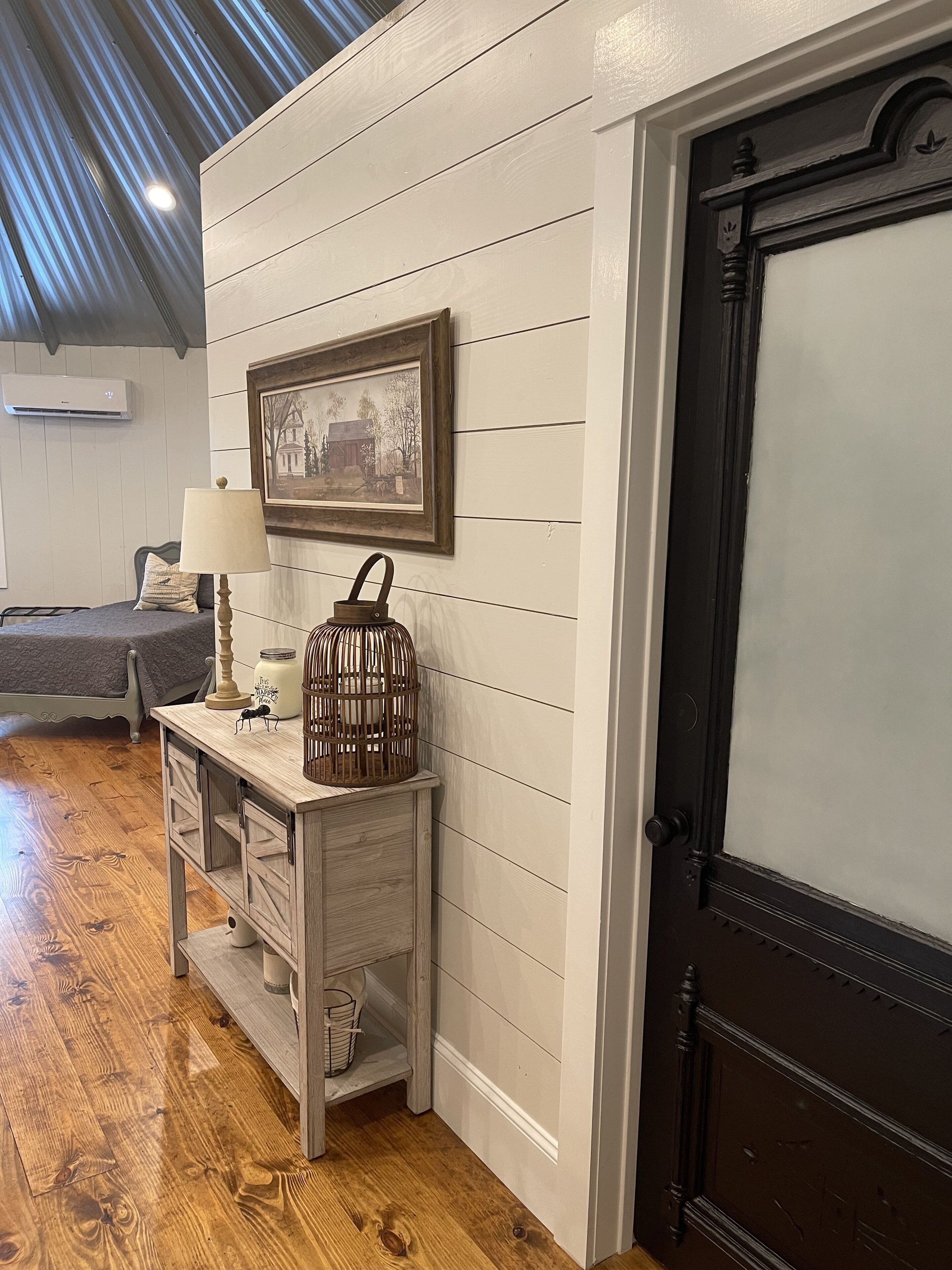 Een kamer met een houten vloer, een witte wand met horizontale planken, een lijst met een afbeelding, een nachtkastje met een lamp, een klein console tafeltje met een kaars en een vogelkooi, en een donkere houten deur.