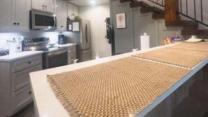 Une cuisine moderne avec des armoires blanches, des appareils en acier inoxydable, un grand îlot central agrémenté d’un set de table en fibres tressées et un escalier avec une rampe en fer forgé.