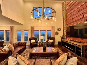 A spacious living room with leather sofas, a wooden coffee table, and a large TV. There are multiple windows offering views of the outdoors.