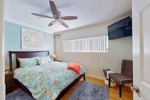 Quarto com cama, ventilador de teto, TV fixada na parede e janela com persianas.