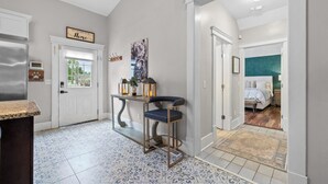Un pasillo con una puerta blanca, una zona de cocina con refrigerador, un taburete de bar y una mesa con objetos decorativos.