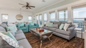 Ein geräumiges Wohnzimmer mit großem Meerblick, Deckenventilator und einer Mischung aus grauen und türkisfarbenen Sofas.