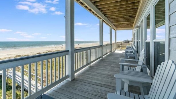 Eine überdachte Holzterrasse mit weißen Stühlen, die auf einen Strand ausgerichtet ist.