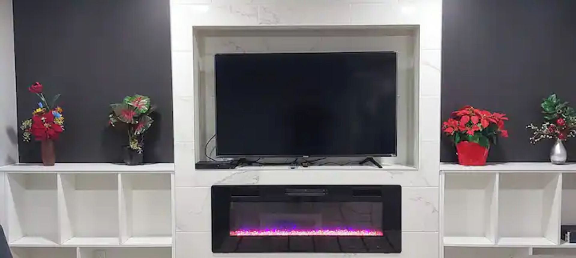 Ett modernt vardagsrum med en platt-TV som är monterad på en vit vägg, och som flankeras av två vita hyllor med krukväxter.