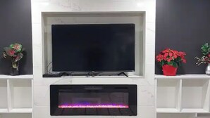 Moderni olohuone, jonka valkoisella seinällä on kiinnitettynä litteänäyttöinen televisio. Seinän molemmin puolin on kaksi valkoista hyllyä, joilla on ruukkukasveja.