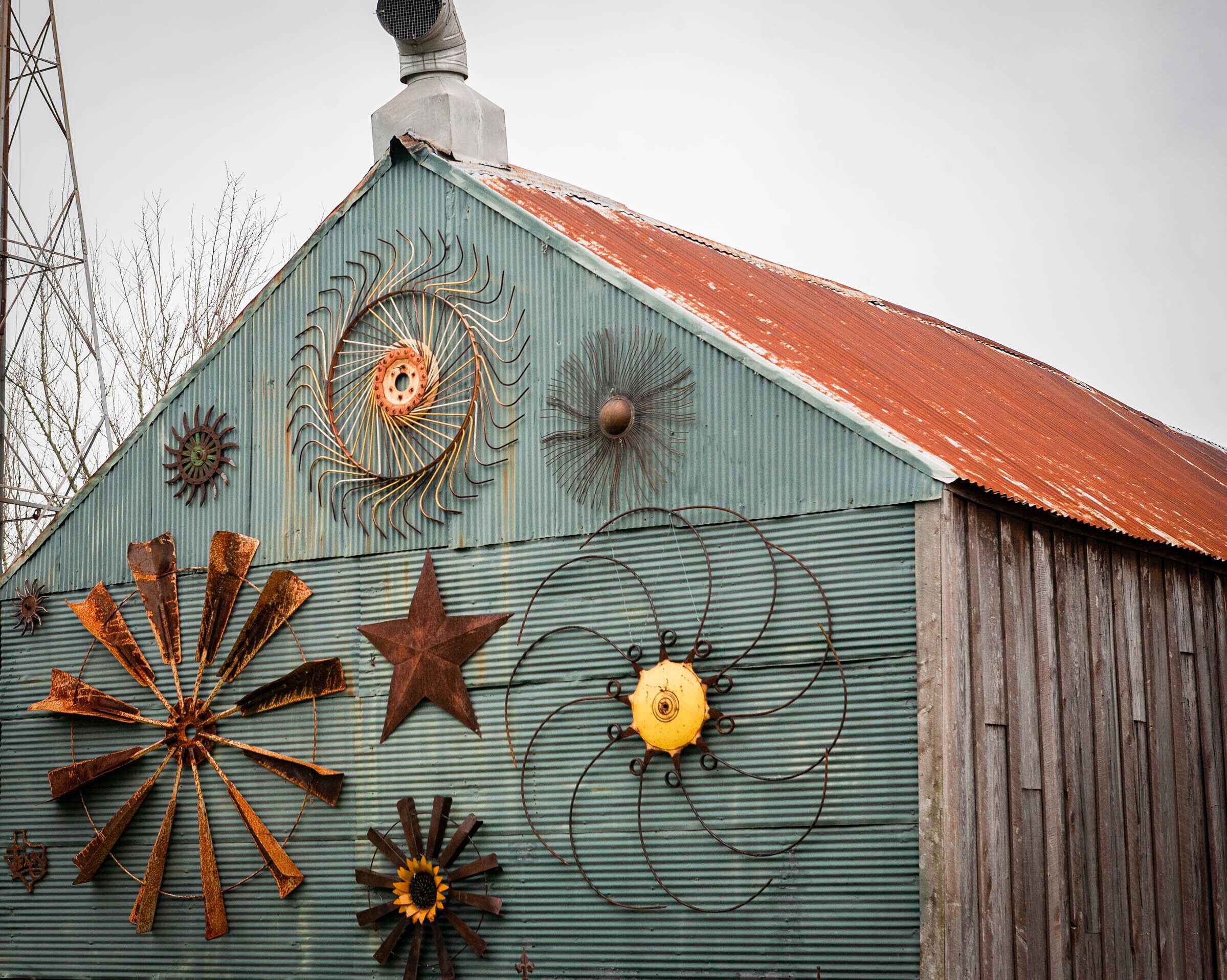 A rustic barn with a corrugated metal exterior, a red roof, and various metal sculptures attached to the wall.