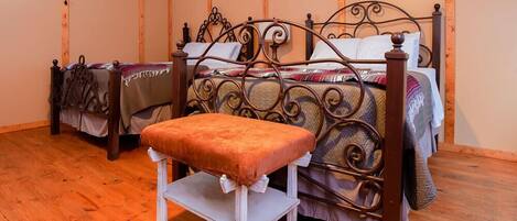 A room with two beds, one with a metal frame and the other with a wooden frame. There is a bench in front of the beds.
