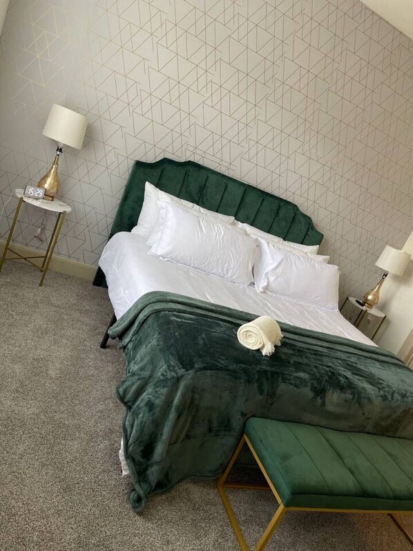 A neatly made bed with a green upholstered headboard and white linens, a green bench at the foot of the bed, and two bedside tables with lamps.