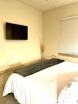 A bedroom with a bed, a flat-screen TV mounted on the wall, a window with blinds, and a vase with dried plants.