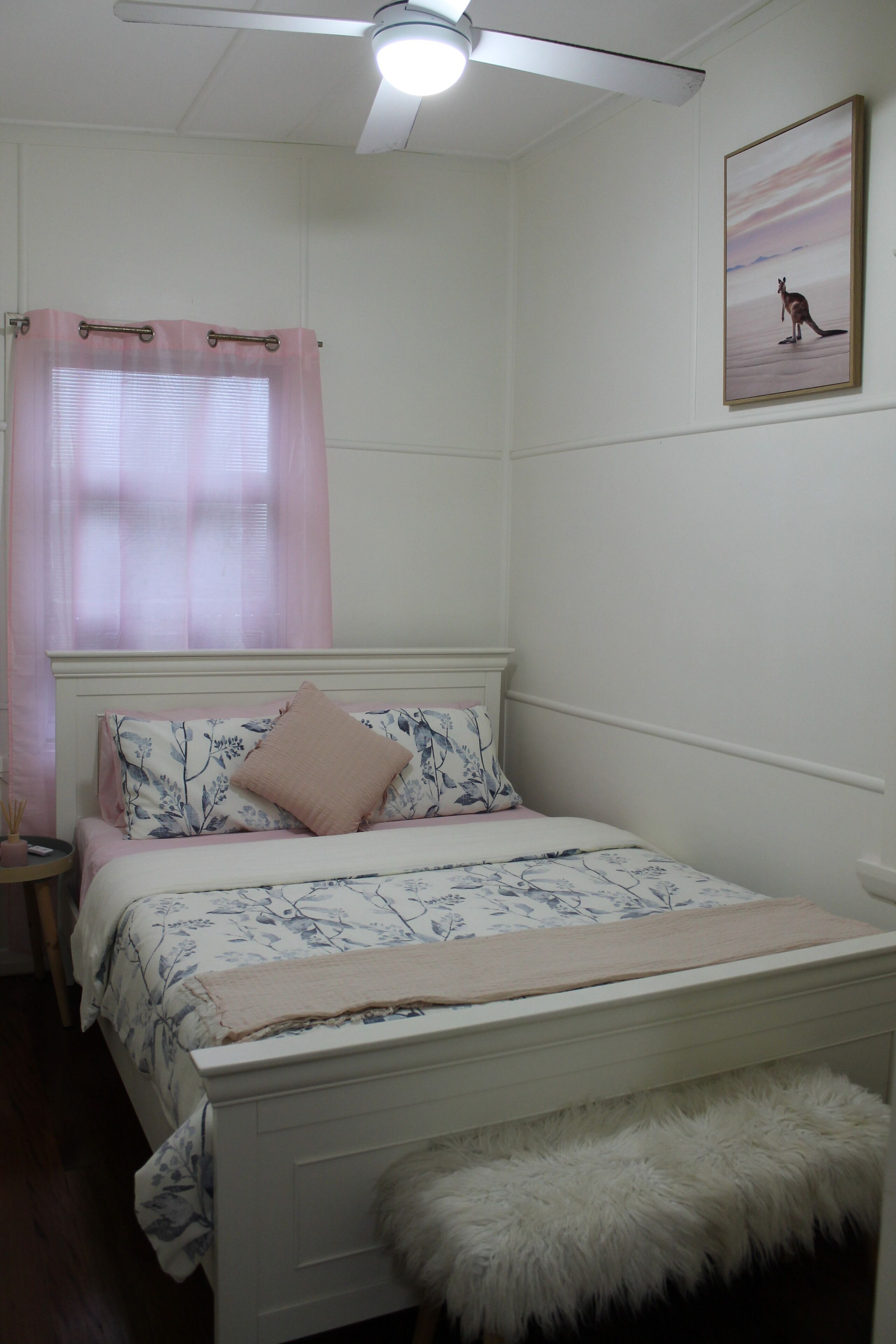 Une chambre à coucher avec un lit, une table de chevet, un banc moelleux blanc, un ventilateur de plafond et une fen être avec des rideaux roses.