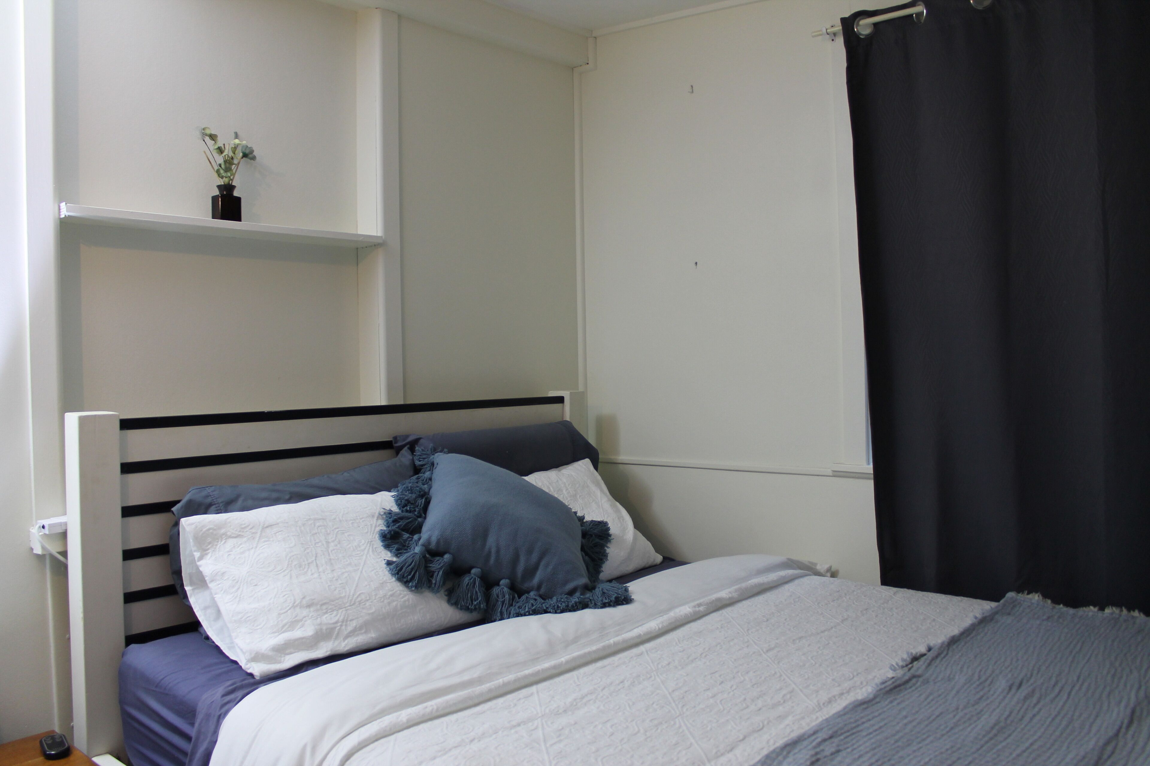Ein Bett mit weißblauer Bettdecke, zwei Kissen und eine kleine Pflanze auf einem weißen Regal darüber.
