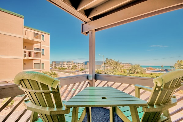 A balcony with two green Adirondack chairs and a small table, overlooking a beach and residential buildings.