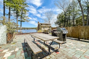 An outdoor patio with a picnic table and grill, surrounded by a wooden fence and trees, overlooking a body of water.