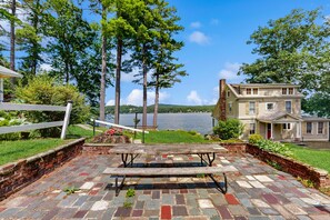 A stone patio with a picnic table, a white fence, and a house with a view of a lake.