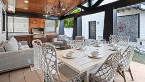 Een terras buiten met een witte eettafel, rieten meubels en een barbecuehoek.