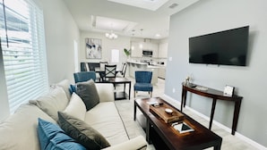 Una sala de estar moderna con sofá, mesa de centro y televisor. Se observa un comedor con sillas y una mesa. La cocina se ve al fondo.