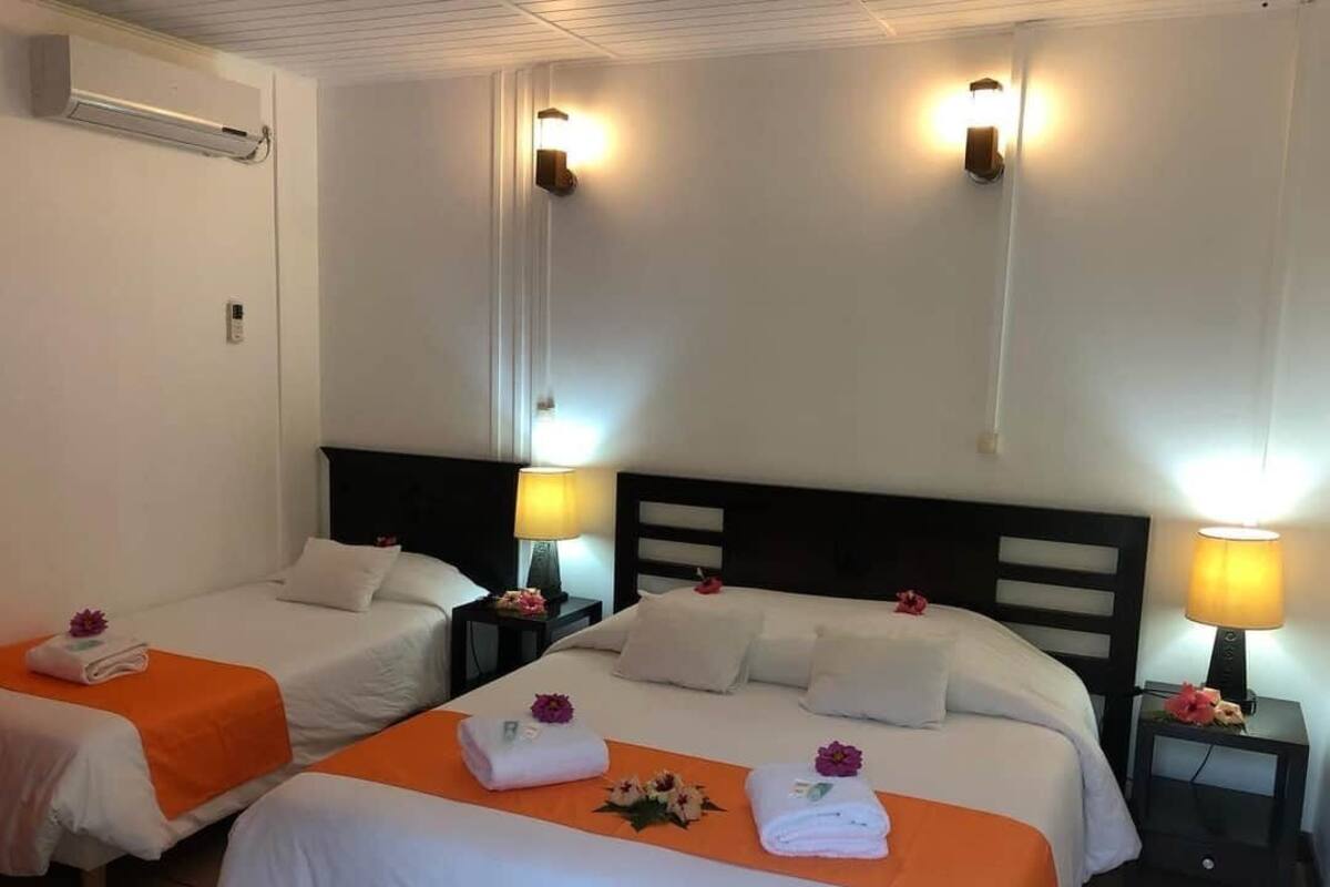 Habitación de hotel con dos camas individuales, cada una con colcha naranja, ropa de cama blanca y flores decorativas. Hay lámparas de noche y un aire acondicionado instalado en la pared.