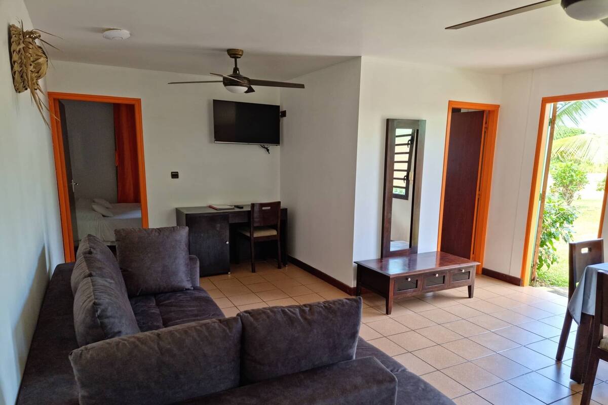 Sala de estar con sofá, televisor montado en la pared, ventilador de techo y escritorio con silla. Hay puertas que dan a espacios exteriores.