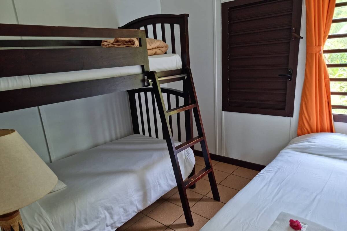 Una habitación con una cama litera, una cama individual, una escalera de madera, una puerta y una ventana con cortinas.